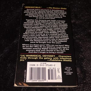 May include: A paperback book with the title "The Hitchhiker's Guide to the Galaxy" is shown. The cover features text and quotes from reviews. The book has a UPC code and ISBN number. The book is a science fiction novel.