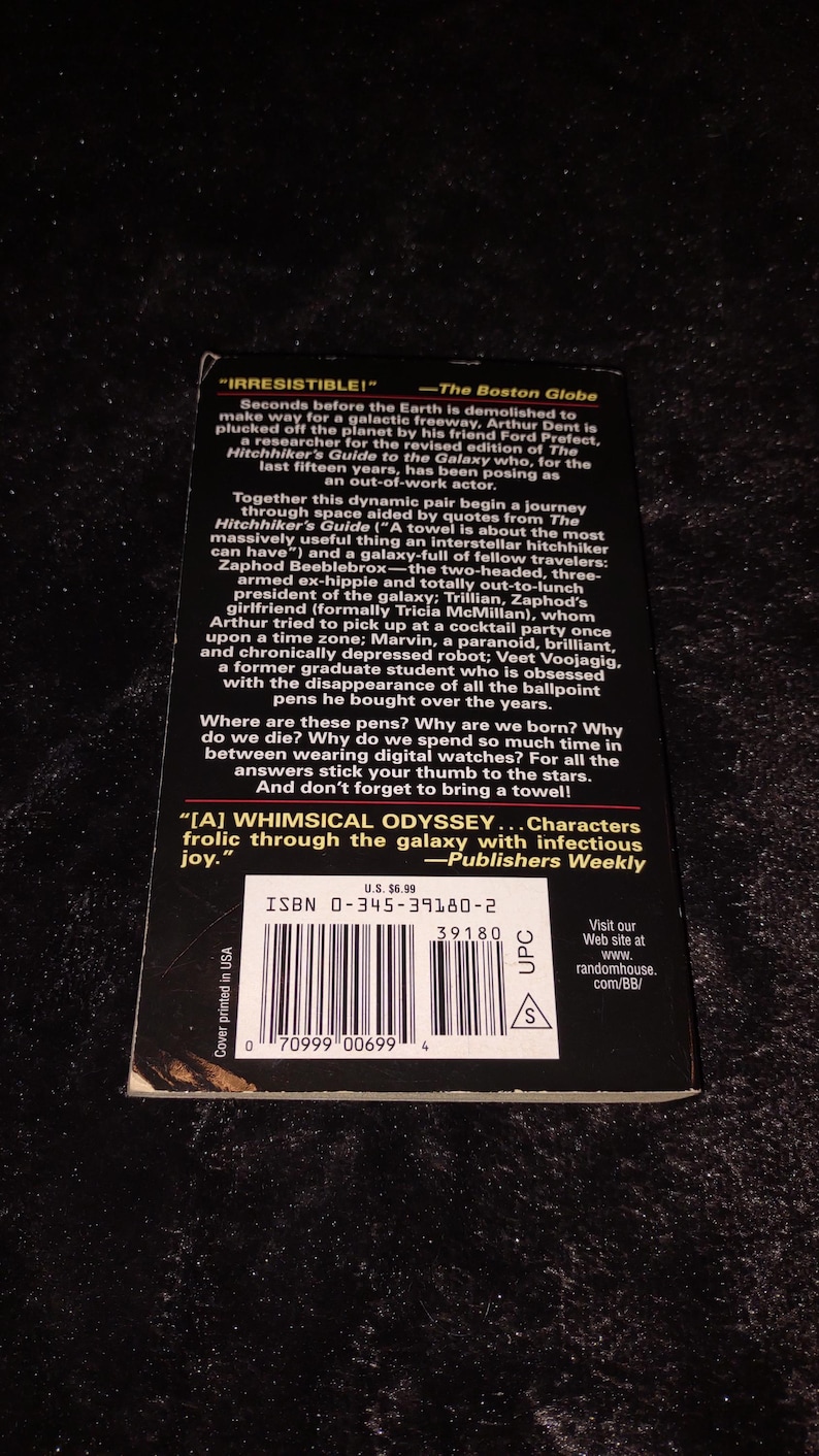 May include: A paperback book with the title "The Hitchhiker's Guide to the Galaxy" is shown. The cover features text and quotes from reviews. The book has a UPC code and ISBN number. The book is a science fiction novel.