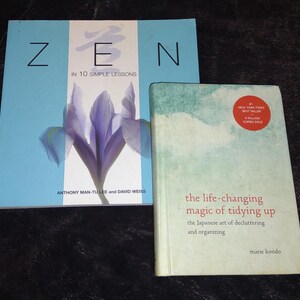 Può includere: Sono esposti due libri. Il primo libro ha una copertina azzurra con la parola "ZEN" a grandi lettere nere e una grafica floreale viola. Il secondo libro ha una copertina bianca con il titolo "the life-changing magic of tidying up".