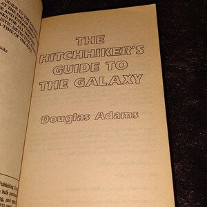 May include: Open book with the title "The Hitchhiker's Guide to the Galaxy" by Douglas Adams. The book's cover is a light tan color with black text. The publisher is Ballantine Books, New York.