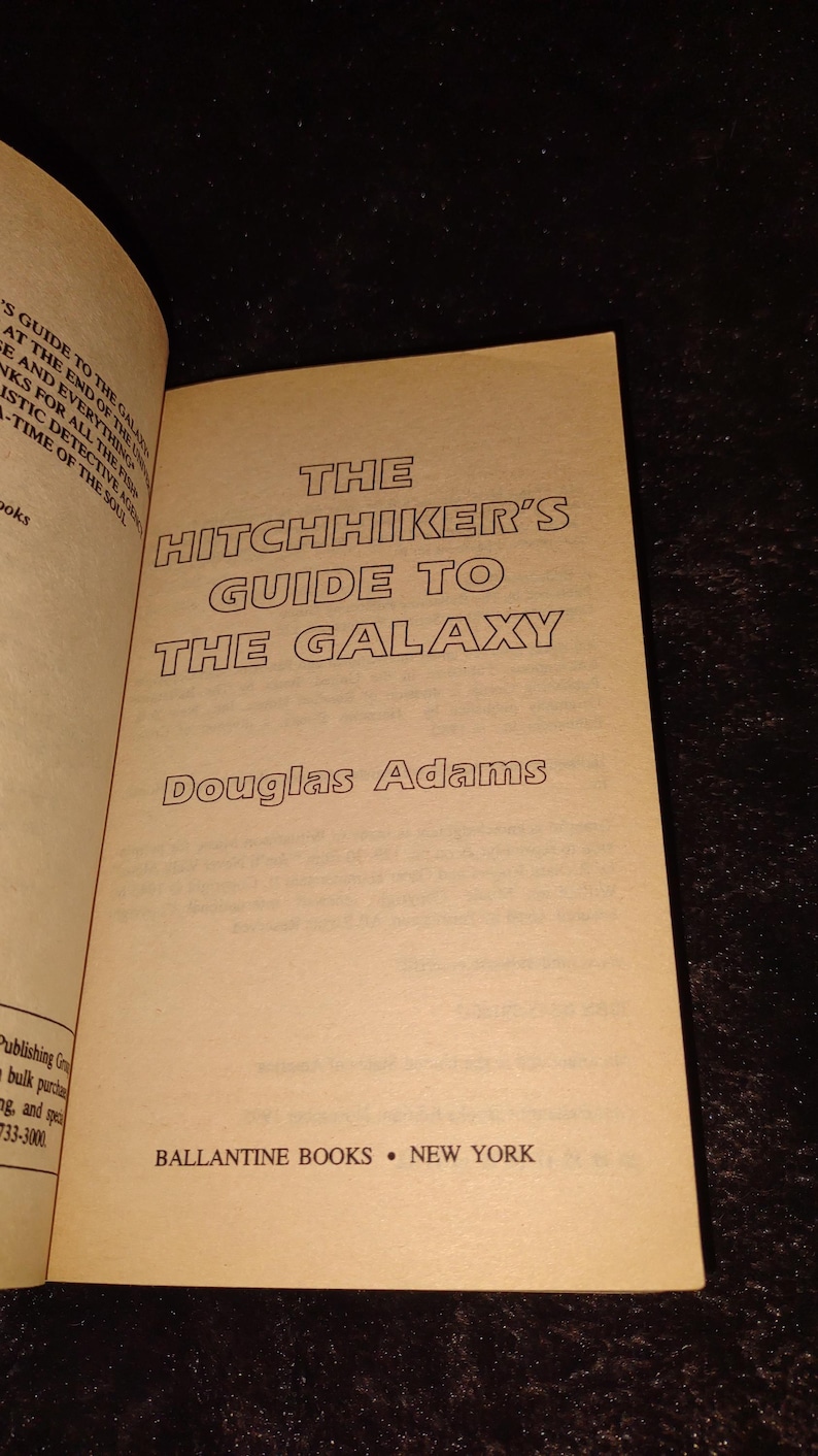 May include: Open book with the title "The Hitchhiker's Guide to the Galaxy" by Douglas Adams. The book's cover is a light tan color with black text. The publisher is Ballantine Books, New York.