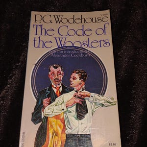 Op de afbeelding: Vintage paperback boek getiteld "The Code of the Woosters" van P.G. Wodehouse. De omslag toont een illustratie van twee mannen. De titel is blauw, met de prijs van $3,95.
