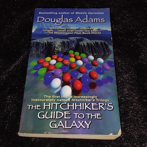 May include: A paperback book titled "The Hitchhiker's Guide to the Galaxy" by Douglas Adams. The cover features colorful, round objects floating over a landscape. The book is part of the Hitchhiker's Trilogy.