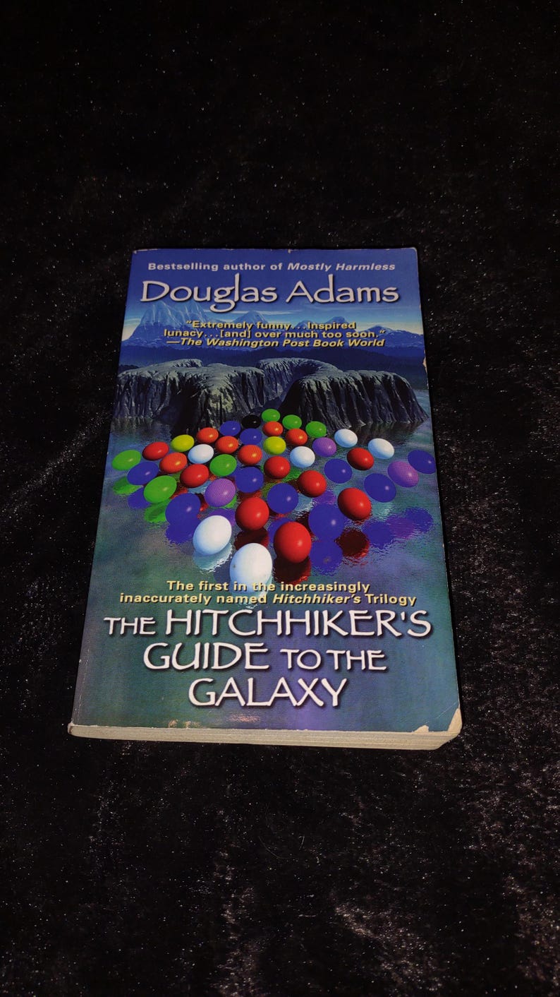 May include: A paperback book titled "The Hitchhiker's Guide to the Galaxy" by Douglas Adams. The cover features colorful, round objects floating over a landscape. The book is part of the Hitchhiker's Trilogy.