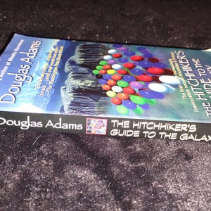 May include: A paperback book titled "The Hitchhiker's Guide to the Galaxy" by Douglas Adams. The cover features a colorful design of various spheres and a blue background. The book is lying on a dark, textured surface.