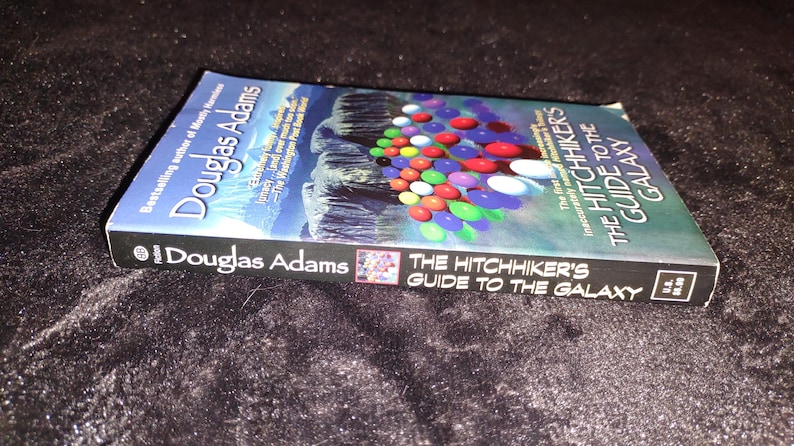 May include: A paperback book titled "The Hitchhiker's Guide to the Galaxy" by Douglas Adams. The cover features a colorful design of various spheres and a blue background. The book is lying on a dark, textured surface.