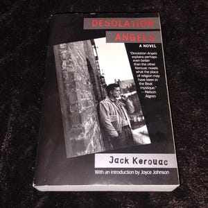 Puede incluir: Un libro de bolsillo titulado "Desolation Angels" de Jack Kerouac. La portada presenta una foto en blanco y negro de un hombre. El título del libro está en letras rojas.