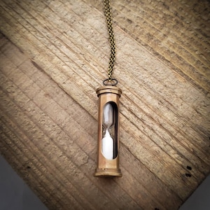 May include: A brass hourglass necklace with a clear glass bulb filled with white sand. The necklace is hanging on a wooden surface.