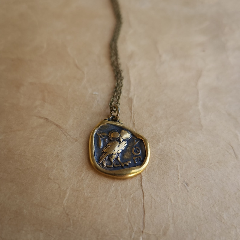 May include: A bronze-toned pendant necklace featuring a detailed relief of an owl. The pendant has a gold-colored border and is suspended from a delicate chain. The owl is depicted with intricate details, and the pendant has Greek lettering.