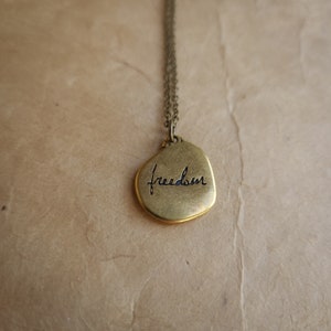 May include: A bronze-colored pendant necklace with the word "freedom" inscribed in a cursive font. The pendant is irregularly shaped and hangs from a delicate chain. The background is a textured, tan surface.