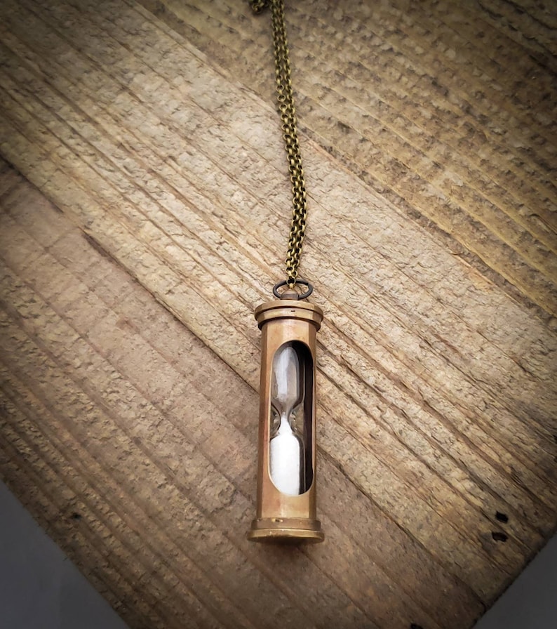 May include: A brass hourglass necklace with a clear glass bulb filled with white sand. The necklace is hanging on a wooden surface.