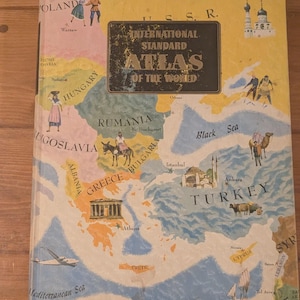 May include: A hardcover book titled International Standard Atlas of the World with a yellow cover featuring a colorful map of Eastern Europe and the Middle East, including illustrations of people, landmarks, and a plane over the Mediterranean Sea.