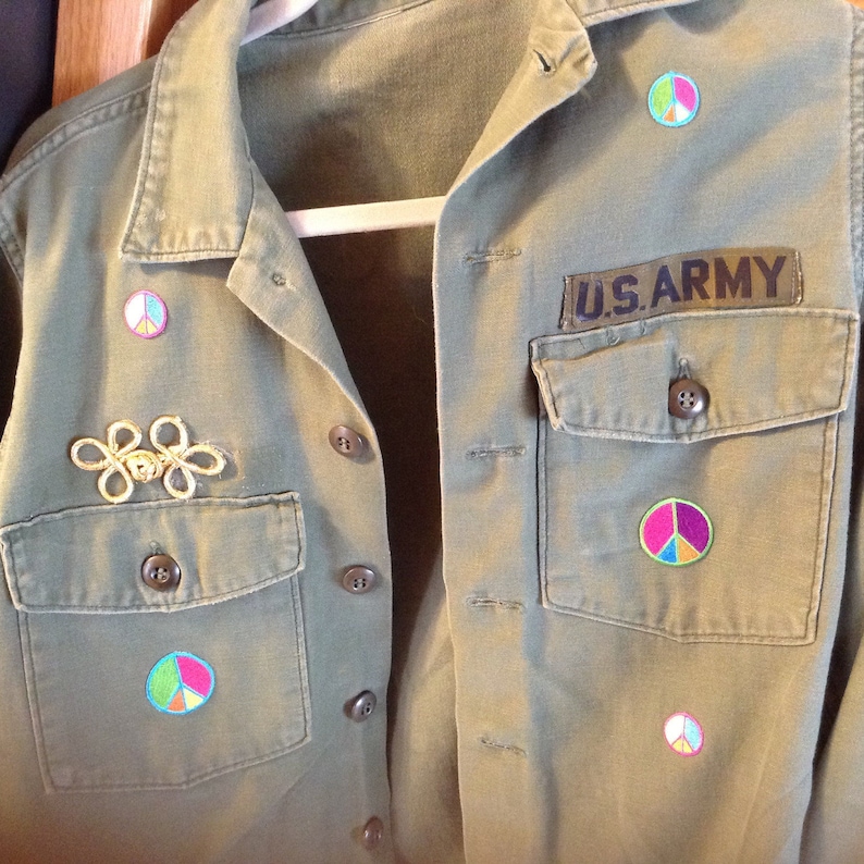 May include: A green military jacket with a U.S. Army patch and several peace sign patches. The jacket has a gold pin with a flower design.