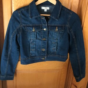 May include: A dark blue denim jacket with a button closure and two front pockets. The jacket has a cropped fit and is made of a soft, comfortable material.