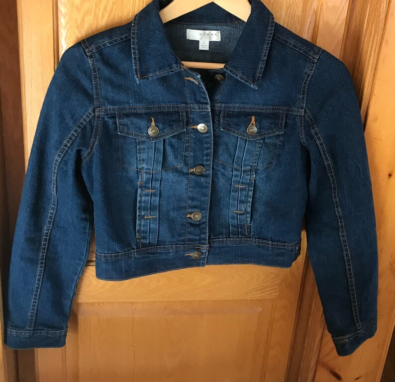 May include: A dark blue denim jacket with a button closure and two front pockets. The jacket has a cropped fit and is made of a soft, comfortable material.
