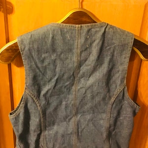 May include: A blue denim vest with a pointed collar and a button closure. The vest is hanging on a wooden hanger.
