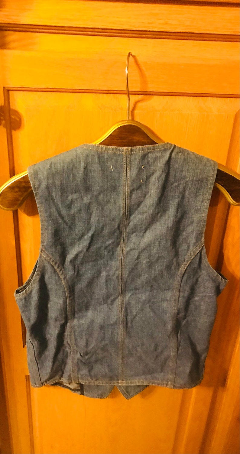 May include: A blue denim vest with a pointed collar and a button closure. The vest is hanging on a wooden hanger.