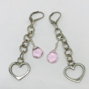 May include: A pair of silver earrings with a delicate chain and a pink teardrop-shaped crystal. Each earring features a heart-shaped charm at the bottom.