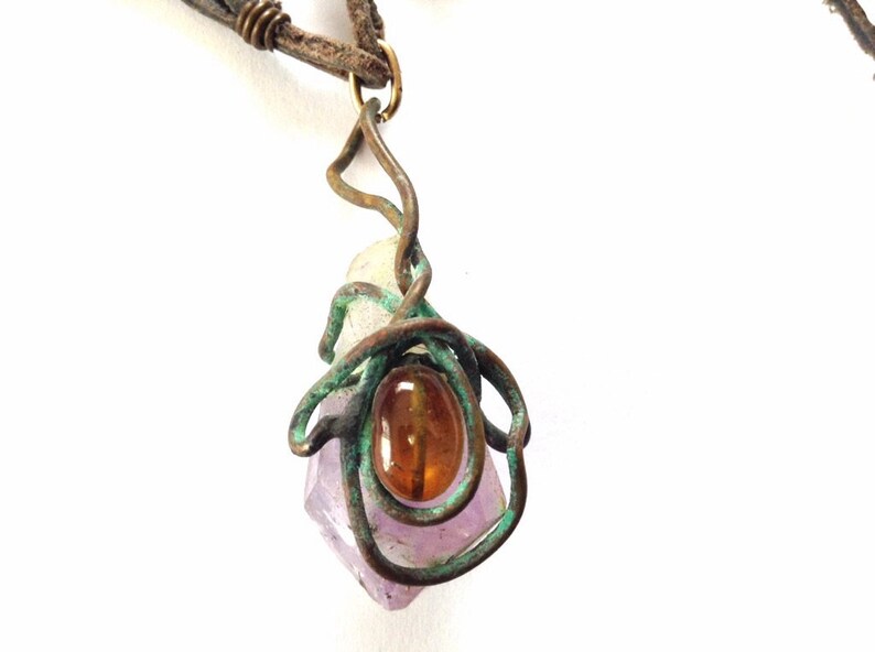 May include: A purple amethyst crystal wrapped in copper wire with a brown gemstone pendant. The pendant is attached to a brown leather cord.
