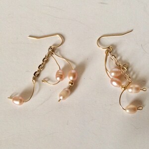May include: A pair of gold dangle earrings with pink pearl accents. The earrings feature a chain and multiple loops with pearls hanging from them.
