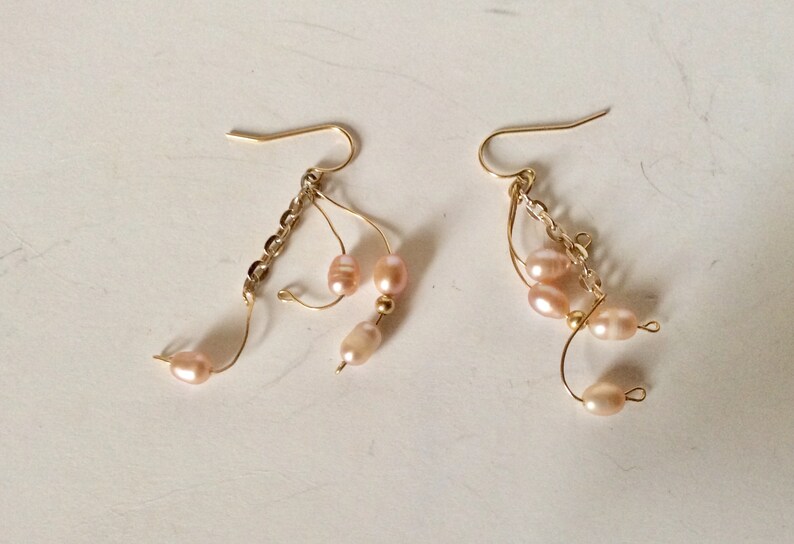 May include: A pair of gold dangle earrings with pink pearl accents. The earrings feature a chain and multiple loops with pearls hanging from them.