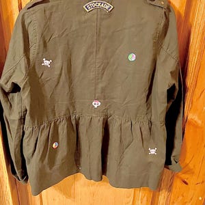 May include: A green utility jacket with a variety of embroidered patches, including a skull and crossbones, a peace sign, and a patch that says "STOCKADE".