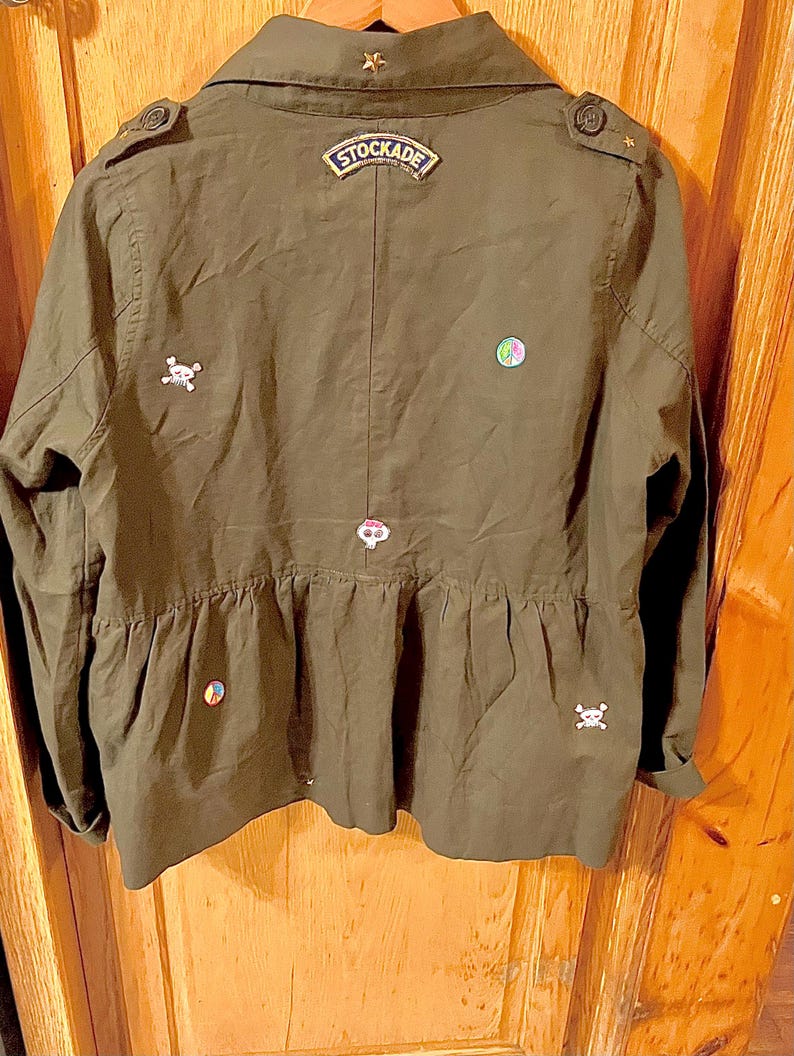 May include: A green utility jacket with a variety of embroidered patches, including a skull and crossbones, a peace sign, and a patch that says "STOCKADE".