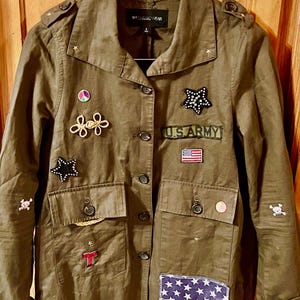 May include: A green utility jacket with various patches and embellishments, including a peace sign, stars, a U.S. Army patch, and a skull and crossbones. The jacket has a button-up front and two flap pockets.