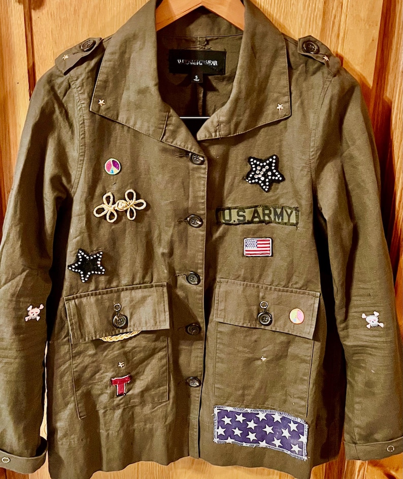 May include: A green utility jacket with various patches and embellishments, including a peace sign, stars, a U.S. Army patch, and a skull and crossbones. The jacket has a button-up front and two flap pockets.