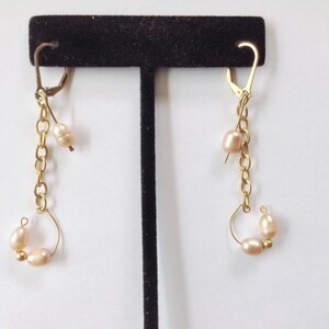 May include: A pair of gold earrings with a chain and multiple small, light pink pearls. The earrings are displayed on a black stand.