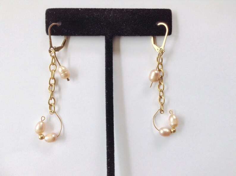 May include: A pair of gold earrings with a chain and multiple small, light pink pearls. The earrings are displayed on a black stand.