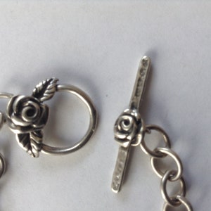 May include: Silver bracelet with a rose-shaped clasp and a toggle closure. The clasp and toggle are both decorated with a rose design.