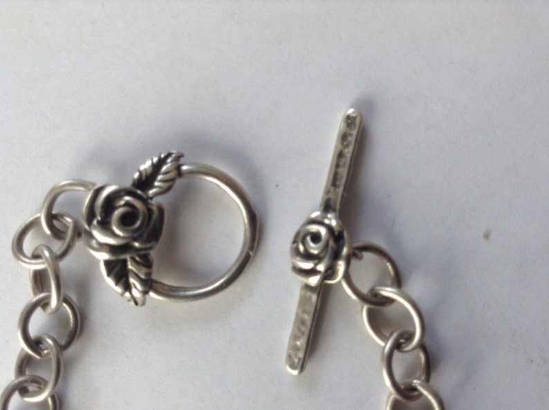 May include: Silver bracelet with a rose-shaped clasp and a toggle closure. The clasp and toggle are both decorated with a rose design.