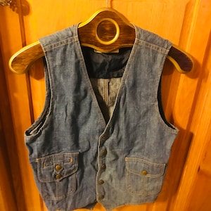 May include: A blue denim vest with two flap pockets and a button closure. The vest is hanging on a wooden hanger.