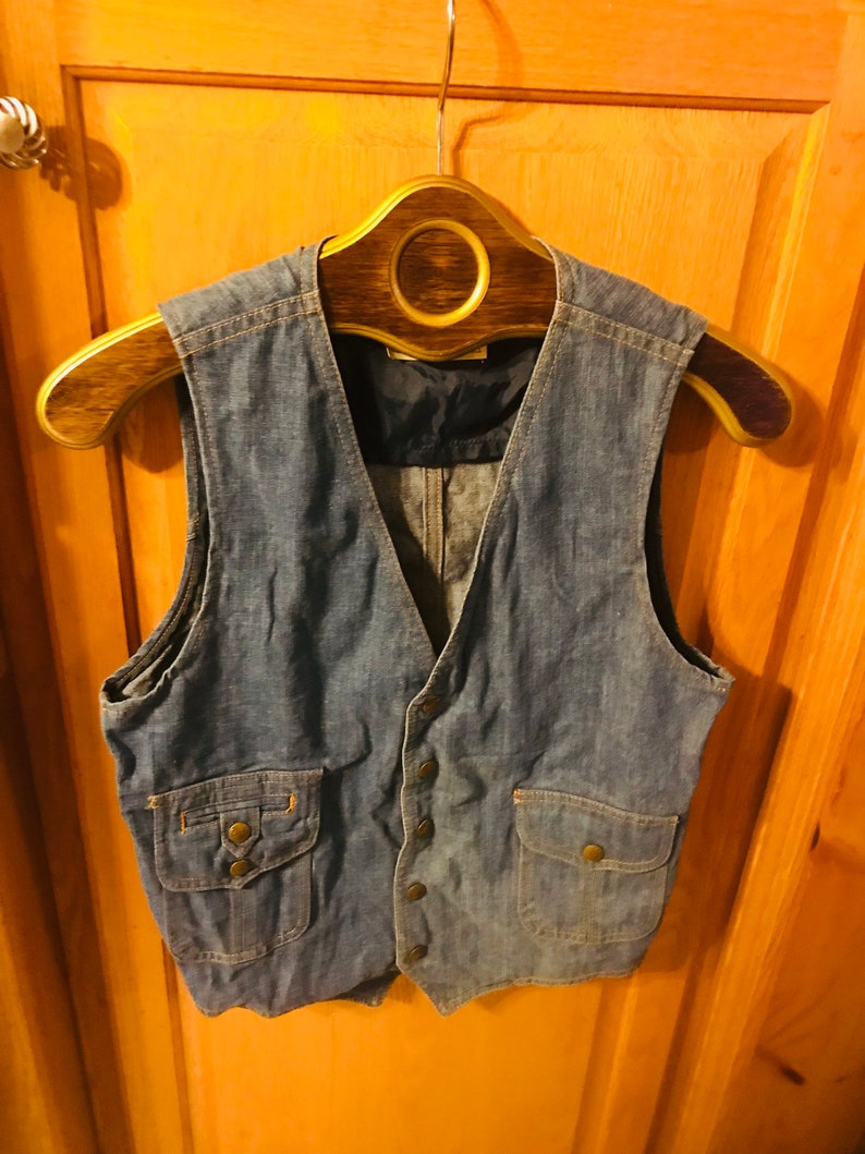 May include: A blue denim vest with two flap pockets and a button closure. The vest is hanging on a wooden hanger.