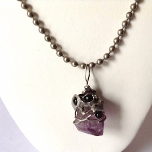 May include: A silver chain necklace with a pendant featuring a large purple amethyst crystal wrapped in silver wire and accented with black beads.