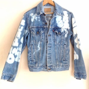 May include: A light blue denim jacket with white bleach splatters and writing on the sleeves. The jacket has a button closure and two pockets on the front.