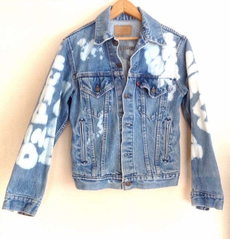 May include: A light blue denim jacket with white bleach splatters and writing on the sleeves. The jacket has a button closure and two pockets on the front.