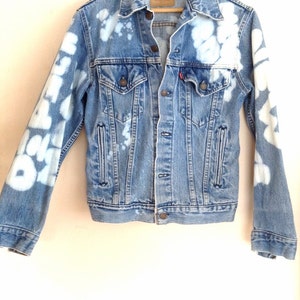 May include: A blue denim jacket with white bleach splatters and designs on the sleeves. The jacket has a button closure and two pockets on the front.