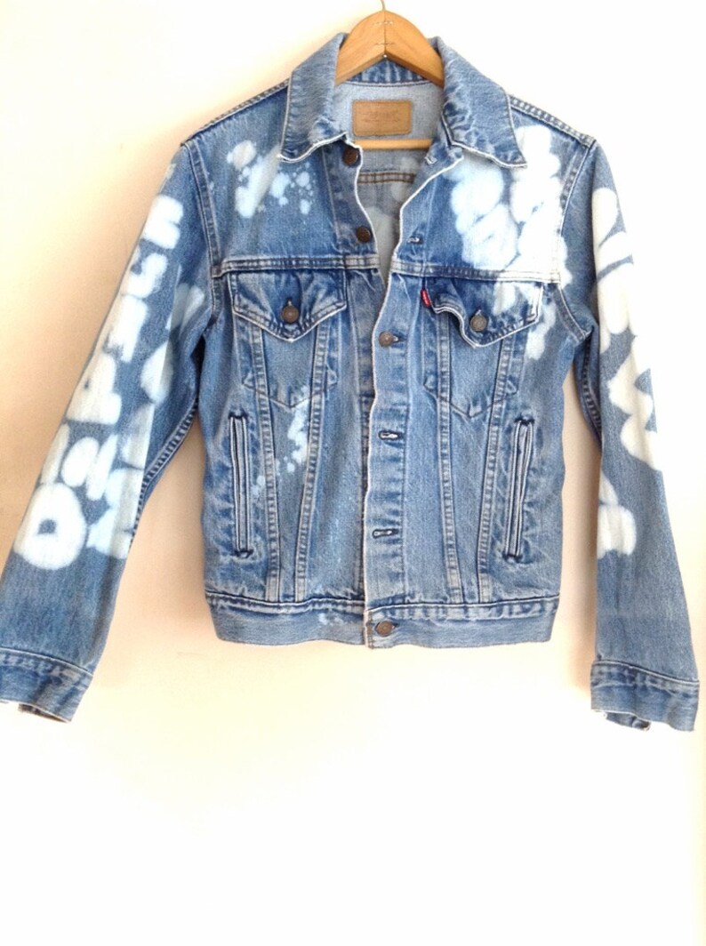 May include: A blue denim jacket with white bleach splatters and designs on the sleeves. The jacket has a button closure and two pockets on the front.