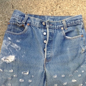 Puede incluir: Vaqueros de mezclilla azul desgastados con múltiples rasgaduras y roturas en todo el tejido. Los vaqueros tienen cierre de botón y cinco bolsillos.