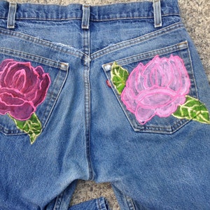 May include: A pair of blue denim jeans with two embroidered roses on the back pockets. The rose on the left pocket is a deep pink and the rose on the right pocket is a light pink.
