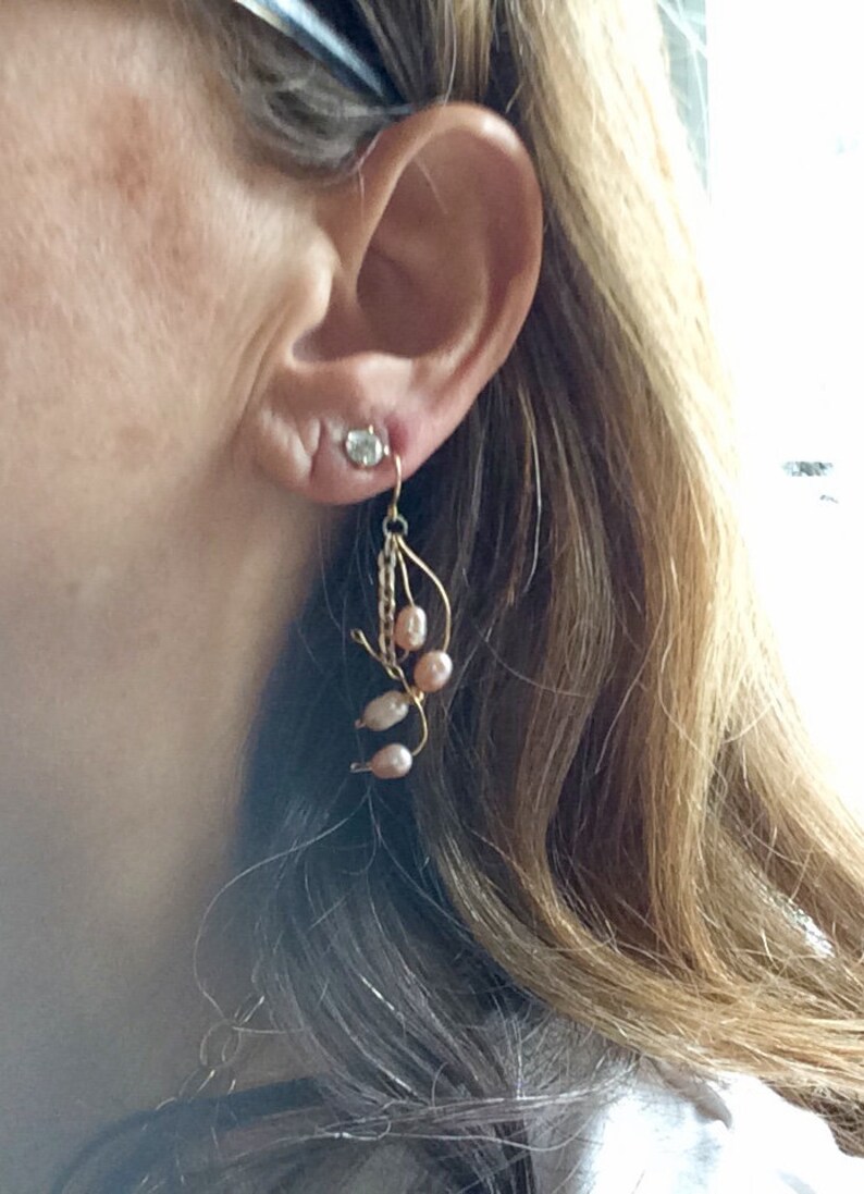 May include: A pair of gold dangle earrings with pink pearls and a clear crystal. The earrings are hanging from a woman's ear.
