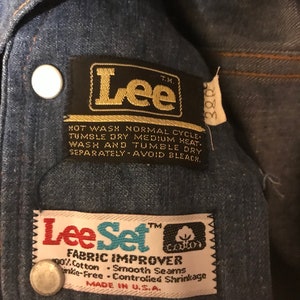 May include: A close-up of a blue denim jacket with two black and gold Lee brand labels. The larger label says "Lee" and has instructions for washing and drying the garment. The smaller label says "Lee Set Fabric Improver" and includes the words "90% Cotton, Smooth Seams, Wrinkle-Free, Controlled Shrinkage, Made in U.S.A."
