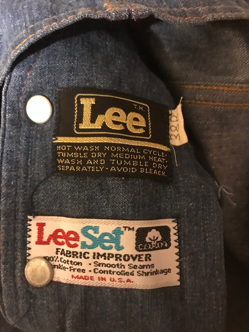 May include: A close-up of a blue denim jacket with two black and gold Lee brand labels. The larger label says "Lee" and has instructions for washing and drying the garment. The smaller label says "Lee Set Fabric Improver" and includes the words "90% Cotton, Smooth Seams, Wrinkle-Free, Controlled Shrinkage, Made in U.S.A."