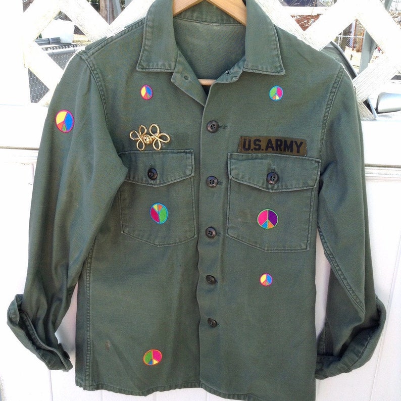 May include: A green military-style shirt with a U.S. Army patch and several colorful peace sign patches. The shirt has a button-up front and two flap pockets.