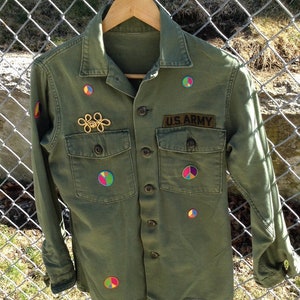 May include: A green US Army utility jacket with a variety of colorful peace sign patches sewn on the front. The jacket has a button-up front and two large pockets.