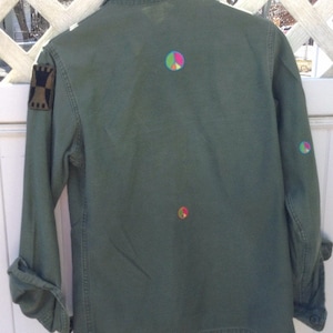 May include: A green military-style jacket with a patch on the left sleeve and three peace signs embroidered on the back.