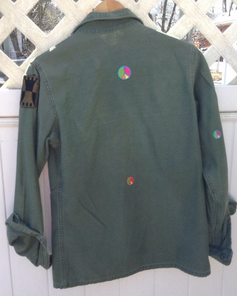 May include: A green military-style jacket with a patch on the left sleeve and three peace signs embroidered on the back.