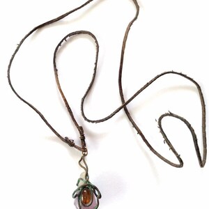May include: A brown leather cord necklace with a pendant featuring a purple and clear crystal wrapped in copper wire. A small orange gemstone is visible in the center of the pendant.